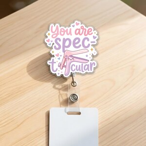 May include: A badge reel with a white rectangular card and a decorative charm. The charm features a pink and purple speculum with the text "You are spectacular" in a whimsical font, surrounded by hearts and stars.