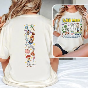 May include: Cream-colored t-shirt featuring a colorful graphic design. The front of the shirt displays the text "LAB WEEK" above a cartoon illustration of lab equipment and characters. The back of the shirt has a vertical design with cartoon characters and lab-related icons.