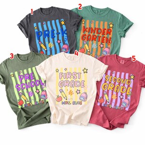 以下が含まれることがあります： 学校をテーマにしたデザインの半袖Tシャツ5枚。各シャツには、「Pre-K」や「First Grade」などの学年が表示され、鉛筆、リンゴ、バックパックなどの学用品が描かれています。シャツの色は、グレー、グリーン、レッドなど様々です。