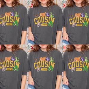 May include: Dark gray t-shirt with the text "COUSIN CREW" in yellow block letters. The shirt features cartoon animal graphics above the text. The shirt is a short-sleeved, crew neck style.