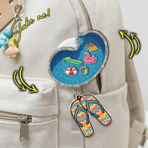May include: A backpack with a pool-shaped keychain featuring a miniature pool scene with a flamingo float, a beach ball, and a life preserver. Below, a pair of colorful flip-flops hangs. The text "Shake me!" is written in a yellow font.