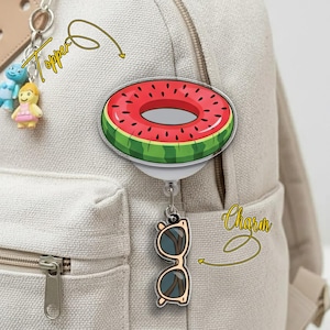 May include: A watermelon-themed badge reel with a red and green design, and a charm of sunglasses. The badge reel is attached to a beige backpack. A small keychain with two figurines is also visible.