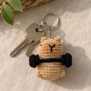 May include: A tan crochet bear keychain with a black barbell. The bear has a black stitched face and is holding the barbell. Two keys are next to the keychain. The keychain is on a silver ring.