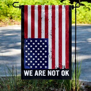 May include: A distressed American flag garden flag with red and white stripes and a blue square with white stars. The flag has the text "WE ARE NOT OK" in white letters on a black background.