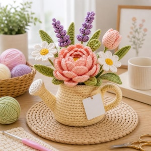 May include: A handmade crochet floral arrangement in a yellow watering can. The arrangement includes a large pink peony, daisies, lavender, and a pink tulip, with green leaves. The watering can sits on a woven mat, with yarn and a crochet hook in the background.