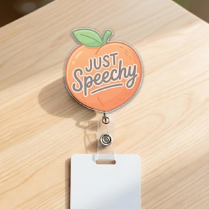 May include: A badge reel featuring a peach design with the words "JUST SPEECHY" in a script font. The peach is orange with a green leaf and a silver outline. The reel has a clear plastic strap and a white rectangular card holder.