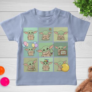 May include: Pale blue baby t-shirt with a grid of nine cartoon images of a green alien character. Each image shows the character in different poses, holding balloons, a cookie, and a yellow ball. The t-shirt is made of soft material.