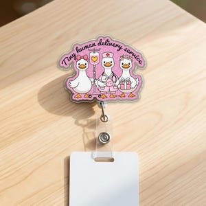 May include: A pink badge reel featuring three cartoon geese. The geese are dressed as a patient, a nurse, and a gift-giver. The text "Tiny human delivery service" is written above the geese in a cursive font. The badge reel has a clear plastic retractable cord and a white card holder.
