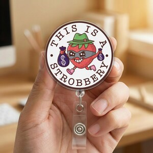 May include: A round badge reel with a cartoon strawberry wearing a green hat and eye mask, holding money bags. The text "THIS IS A STROBBERY" is printed around the image. The reel has a clear plastic clip and a retractable cord.