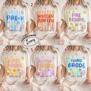 以下が含まれることがあります： アイボリー色のTシャツ。各シャツには、「Pre-K」や「Kindergarten」などの学年レベルがカラフルな文字で表示され、鉛筆、リンゴ、バックパックなどの学校をテーマにしたグラフィックが描かれています。「Mrs. Alice」の文字も含まれています。