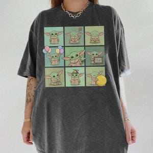 May include: Dark grey t-shirt with a grid of nine cartoon images of a green alien character. Each image shows the character in different poses, some holding objects like balloons and a ball. The shirt has a relaxed fit.