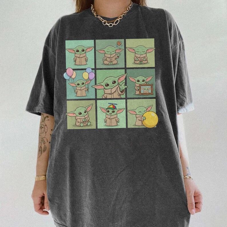 May include: Dark grey t-shirt with a grid of nine cartoon images of a green alien character. Each image shows the character in different poses, some holding objects like balloons and a ball. The shirt has a relaxed fit.
