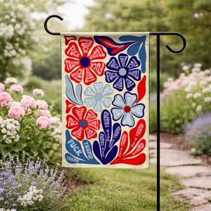 May include: A garden flag with a floral design. The flowers are red, white, and blue, with the words "FUCK TRUMP" in the center of each flower. The flag is hanging on a black metal stand in a garden setting.