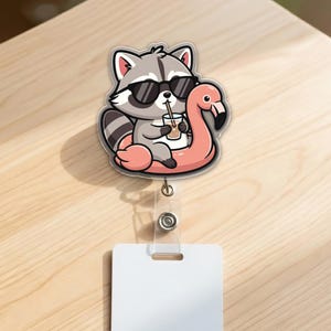 May include: A fun, cartoon-style badge reel featuring a raccoon wearing sunglasses and floating on a pink flamingo pool float. The raccoon is holding a drink with a straw. The reel has a clear plastic strap and a white rectangular tag.