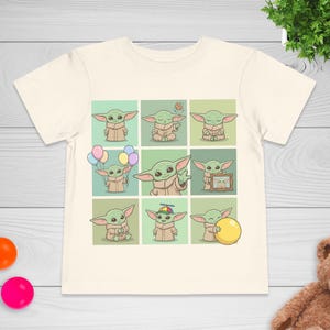 May include: Cream-coloured t-shirt featuring a grid of nine cartoon images of a green alien character. The character is depicted in various poses, holding balloons, a cookie, and a yellow ball. The shirt is made of soft material.