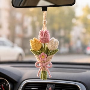 May include: A handmade crochet bouquet of tulips in a car. The bouquet features a pink, yellow, and white tulip, with green leaves and a pink bow. The car accessory hangs from the rearview mirror.