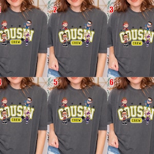 May include: Dark gray t-shirt featuring the text "Cousin Crew" in a yellow and white design, with cartoon characters above and below the text. The shirt has short sleeves and a crew neck.