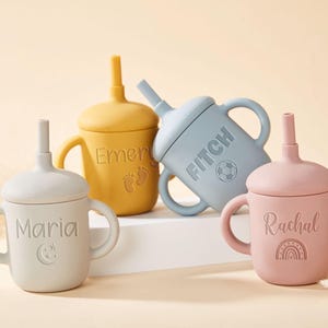 May include: Four silicone sippy cups in pastel colors: gray, yellow, blue, and pink. Each cup has a lid, handles, and a straw. The cups are personalized with names and symbols: Maria, Emery, Fitch, and Rachael. A baby product.
