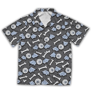 Puede incluir: Una camisa de manga corta con fondo gris y un patrón repetido de huesos blancos, alas de murciélago azules y globos oculares. La camisa tiene un cuello clásico y un corte relajado.