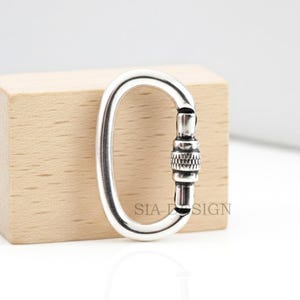 May include: A silver-colored oval carabiner with a screw lock mechanism. The carabiner is positioned in front of a small, light-colored wooden block. The text "SIA DESIGN" is printed on the carabiner.