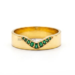 Könnte beinhalten: Ein goldener Ring mit einem V-förmigen Ausschnitt in der Mitte, der mit einer Reihe kleiner, runder, leuchtend grüner Edelsteine besetzt ist. Das Wort SIA DESIGN ist in schwarzer Schrift auf der glatten Goldoberfläche des Rings eingraviert.