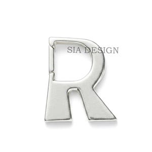 Pode incluir: Um berloque prateado com a letra "R" e um fecho, sobre um fundo branco. O texto "SIA DESIGN" está impresso no canto superior direito da letra. Este berloque é provavelmente para joias ou projetos de artesanato.