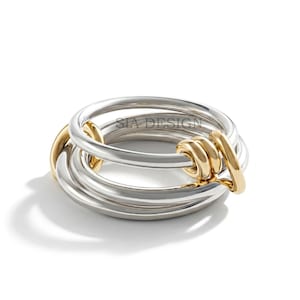 Peut inclure: Une bague multi-anneaux composée de plusieurs anneaux argentés entrelacés et d'un anneau doré. Le texte SIA DESIGN est clairement visible sur la surface de l'anneau argenté supérieur. Le design présente des boucles en métal lisse et poli.