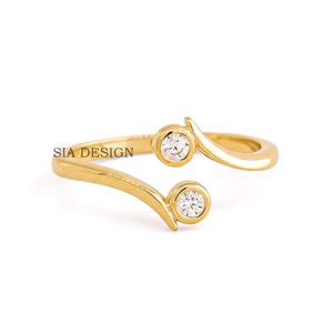 May include: A gold-colored ring with two clear, round gemstones set in bezel settings. The ring has a unique, open design with curved bands. The text "SIA DESIGN" is visible on the ring.
