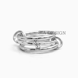 以下が含まれることがあります： ユニークなデザインのシルバーリング。リングは、円形のリンクで接続された複数のバンドが特徴です。「SIA DESIGN」の文字がリングに表示されています。リングは白い背景に表示されています。