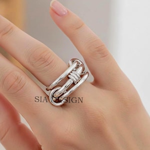 May include: A silver ring with a distinctive, linked design. The ring showcases a series of interconnected, elongated loops and a central, wrapped detail. The text "SIA DESIGN" is visible on the ring.