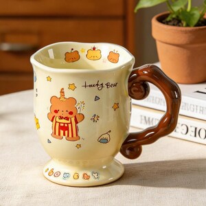 Könnte beinhalten: Eine cremefarbene Keramiktasse mit braunem Henkel und Fuß. Die Tasse ist mit einem Cartoon-Bären, Sternen und dem Text "Lucky Bear" verziert.