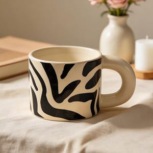 Puede incluir: Una taza de cerámica color crema con un diseño de rayas de cebra negras. La taza tiene un asa redondeada y está sobre un mantel beige. Un libro, un jarrón con rosas rosas y una vela están en el fondo.