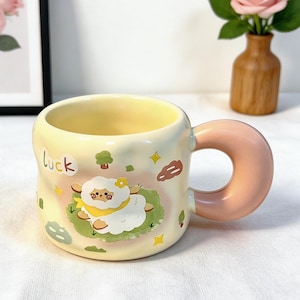 May include: A ceramic mug with a light yellow exterior and a pink handle. The mug features a cartoon sheep design with the word "luck" written on the side. The interior of the mug is yellow.