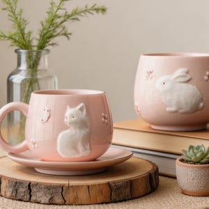 May include: A pink ceramic mug with a white cat design, saucer, and a matching pink ceramic planter with a white bunny design. The mug and planter are decorated with floral accents. A small succulent in a terracotta pot is also present.