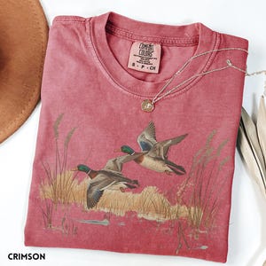 May include: Crimson-colored t-shirt featuring a graphic of two ducks in flight over a marsh. The shirt has a crew neck and short sleeves. A silver necklace with a pendant is draped across the shirt. The shirt is labeled "Comfort Colors" and is size S-P-CH.