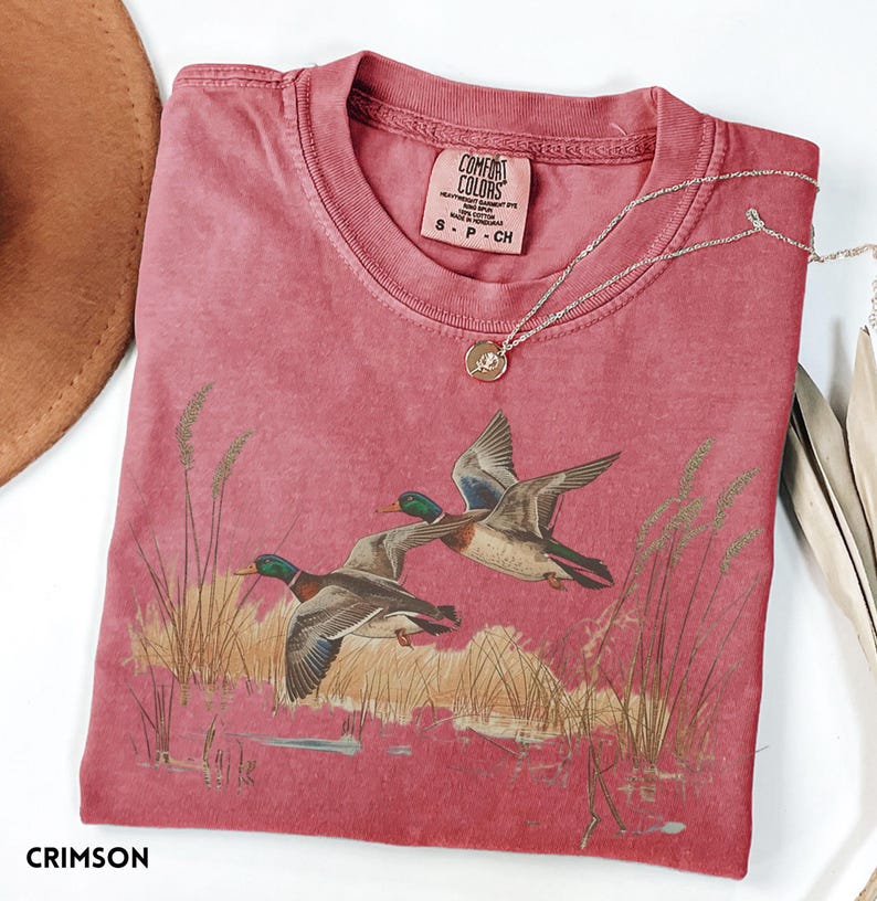 May include: Crimson-colored t-shirt featuring a graphic of two ducks in flight over a marsh. The shirt has a crew neck and short sleeves. A silver necklace with a pendant is draped across the shirt. The shirt is labeled "Comfort Colors" and is size S-P-CH.