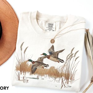 May include: Ivory-colored t-shirt featuring a nature scene with two ducks in flight over a marsh. The shirt has a crew neck and short sleeves. A gold necklace with a pendant rests on the shirt. The word "IVORY" is printed at the bottom.