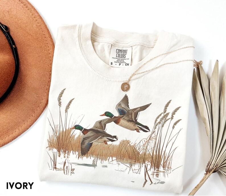 May include: Ivory-colored t-shirt featuring a nature scene with two ducks in flight over a marsh. The shirt has a crew neck and short sleeves. A gold necklace with a pendant rests on the shirt. The word "IVORY" is printed at the bottom.
