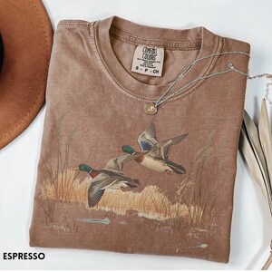 May include: Espresso-colored t-shirt featuring a graphic of two ducks in flight over a marsh. The shirt has a crew neck and short sleeves. A silver necklace with a pendant rests on the shirt. The brand label "Comfort Colors" is visible.