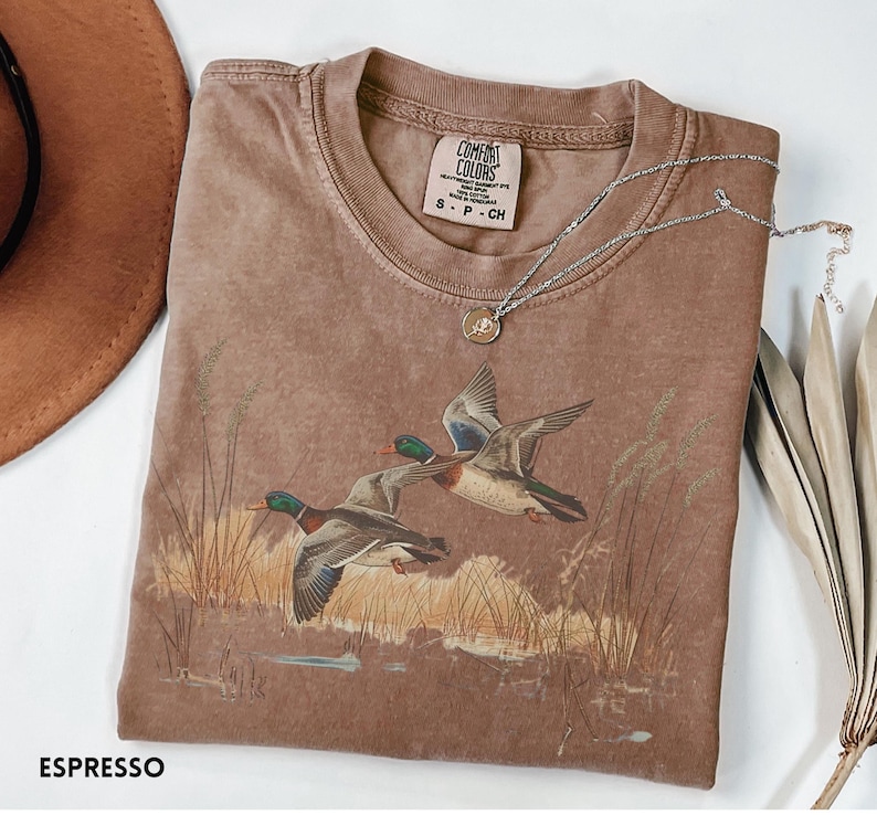 May include: Espresso-colored t-shirt featuring a graphic of two ducks in flight over a marsh. The shirt has a crew neck and short sleeves. A silver necklace with a pendant rests on the shirt. The brand label "Comfort Colors" is visible.