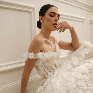 Op de afbeelding: Een witte trouwjurk met een geplooide off-the-shoulder halslijn en een volle rok versierd met bloemenapplicaties en glinsterende versieringen. De jurk heeft een getailleerd lijfje en een vloeiende, gelaagde rok, wat zorgt voor een romantische en elegante uitstraling.