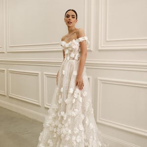 Op de afbeelding: Een witte off-the-shoulder trouwjurk met bloemenversieringen. De jurk heeft een nauwsluitend lijfje en een zwierige rok, versierd met 3D bloemenapplicaties. De jurk is gemaakt van een glinsterende stof, wat een romantische en elegante uitstraling cre&euml;ert.