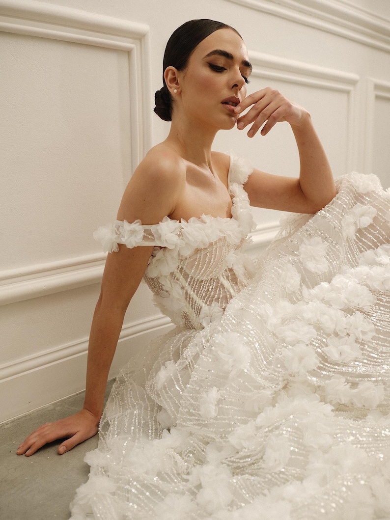 Op de afbeelding: Een witte trouwjurk met een geplooide off-the-shoulder halslijn en een volle rok versierd met bloemenapplicaties en glinsterende versieringen. De jurk heeft een getailleerd lijfje en een vloeiende, gelaagde rok, wat zorgt voor een romantische en elegante uitstraling.