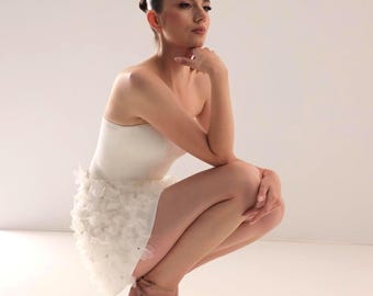 May include: A woman in a white strapless top and a white skirt with floral details, wearing beige pointed heels. She is squatting, with her hand on her chin. The background is plain white.