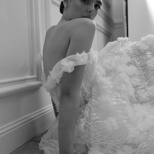 Op de afbeelding: Een zwart-witfoto van een vrouw in een witte off-the-shoulder jurk. De jurk heeft ingewikkelde bloemenversieringen en een zwierige rok. De vrouw kijkt over haar schouder, met haar haar opgestoken.