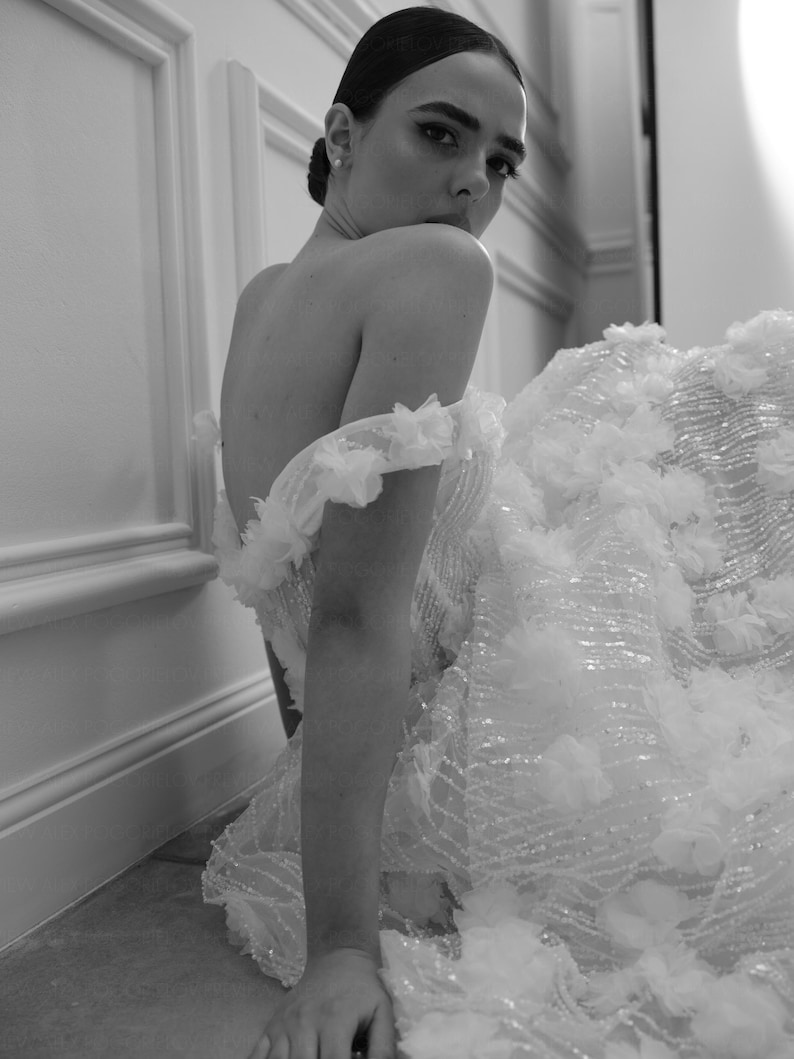 Op de afbeelding: Een zwart-witfoto van een vrouw in een witte off-the-shoulder jurk. De jurk heeft ingewikkelde bloemenversieringen en een zwierige rok. De vrouw kijkt over haar schouder, met haar haar opgestoken.