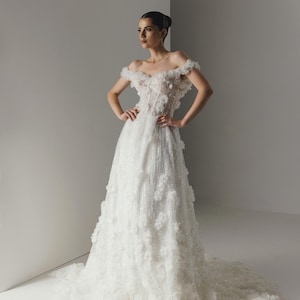 Op de afbeelding: Een witte off-the-shoulder trouwjurk met bloemversieringen. De jurk heeft een nauwsluitend lijfje, een zwierige rok en een lange sleep. De jurk is versierd met delicate bloemenapplicaties en sprankelende details, wat een romantische en elegante uitstraling cre&euml;ert.