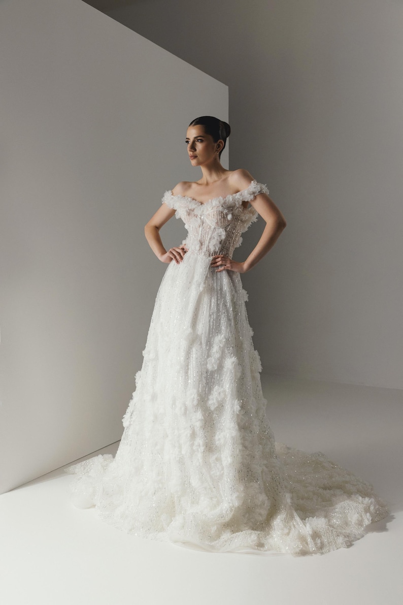 Op de afbeelding: Een witte off-the-shoulder trouwjurk met bloemversieringen. De jurk heeft een nauwsluitend lijfje, een zwierige rok en een lange sleep. De jurk is versierd met delicate bloemenapplicaties en sprankelende details, wat een romantische en elegante uitstraling cre&euml;ert.