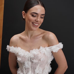 Op de afbeelding: Een witte off-the-shoulder trouwjurk met bloemversieringen. Het lijfje heeft delicate witte bloemen en sprankelende details, met een zwierige rok. De jurk is ontworpen in een romantische en elegante stijl, perfect voor een speciale gelegenheid.