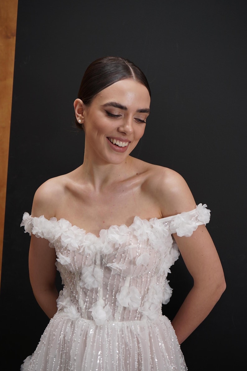 Op de afbeelding: Een witte off-the-shoulder trouwjurk met bloemversieringen. Het lijfje heeft delicate witte bloemen en sprankelende details, met een zwierige rok. De jurk is ontworpen in een romantische en elegante stijl, perfect voor een speciale gelegenheid.